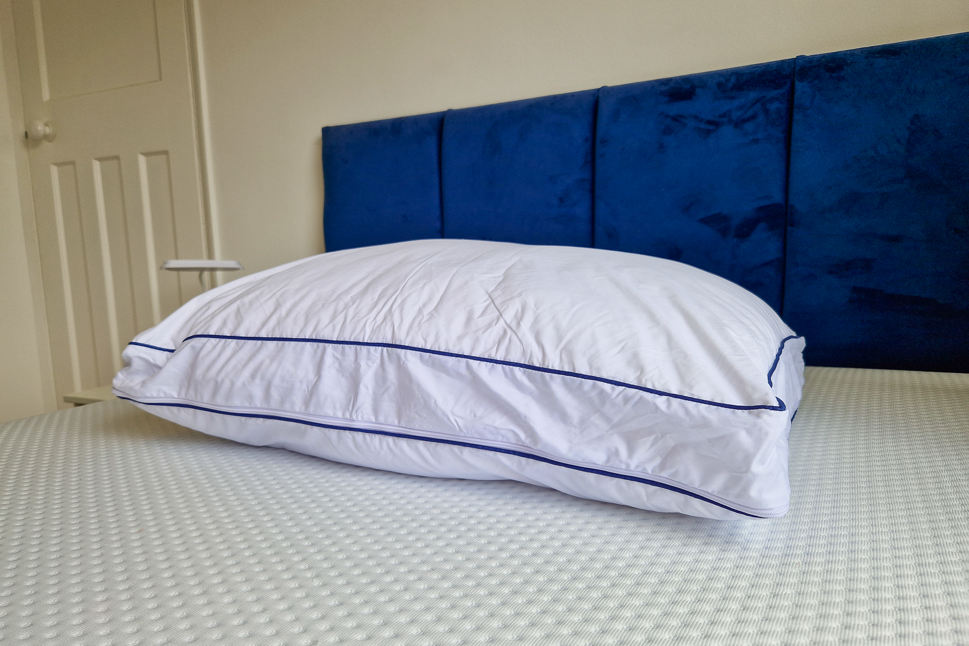 Almofada com fronha branca e linha azul pousada sobre uma cama com cabeceira azul escura.