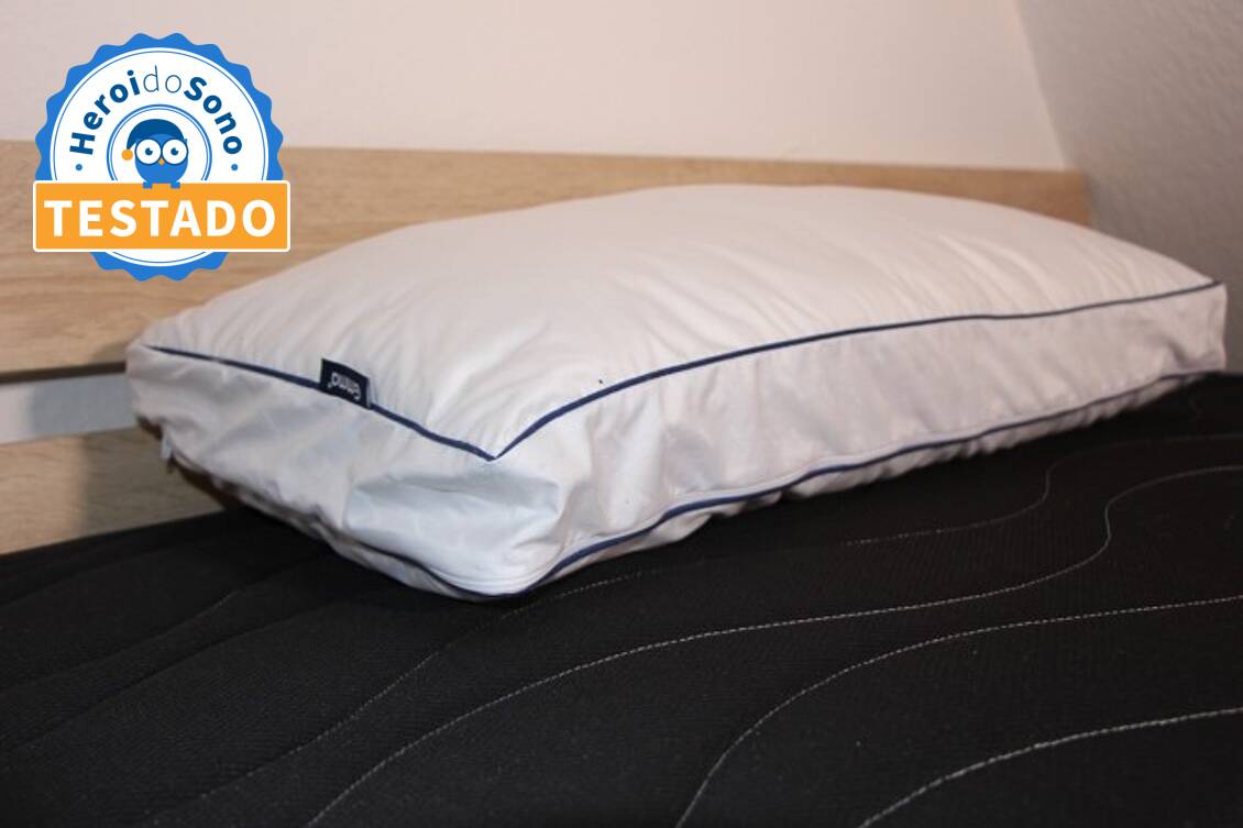 Almofada de microfibra da marca Emma com etiqueta visível, pousada sobre uma cama com lençol escuro e cabeceira de madeira clara.