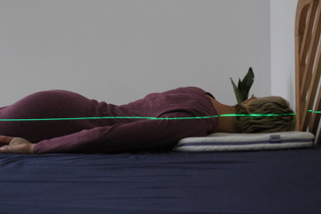 Pessoa de cabelo curto claro deitada de barriga para baixo numa almofada branca com um laser verde a mostrar o alinhamento da coluna.